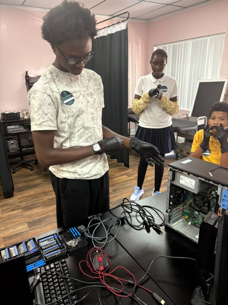 Three young students put on black gloves at a workstation with open computer hardware, screwdriver kits, and cable components